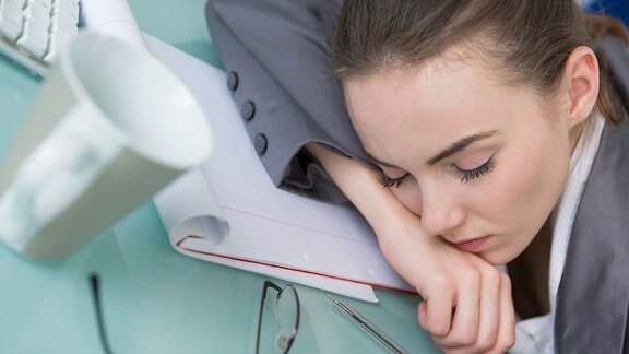 Frau schläft im Büro auf dem Tisch, neben ihr eine Computertastatur, ein Stift und ein Kaffeebecher