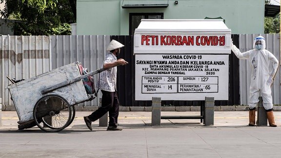 Ein Mann fährt in Jakarta mit seinem Wagen an einem nachgebildeten Sarg vorbei, neben dem ein Schutzanzug tragender Mann steht