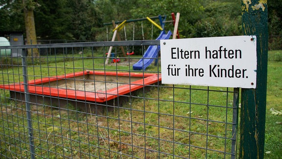 Schild "Eltern haften für ihre Kinder" an einem Zaun vor einem Spielplatz