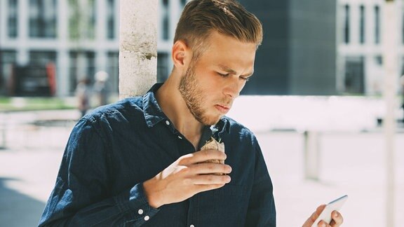 Ein Mann isst im Stehen und schaut auf sein Handy