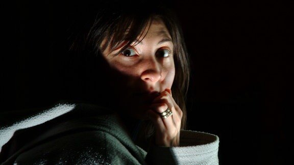 Eine Frau hält sich im Dunkeln die Hand vor den Mund und schaut erschreckt in die Kamera.