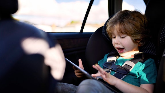 Junge spielt auf dem Rücksitz im Kindersitz auf einem Tablet (Symbolbild)
