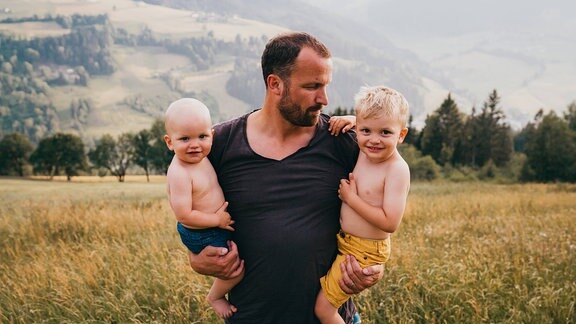 Vater mit zwei Kindern auf dem Arm vor Alm-Kulisse