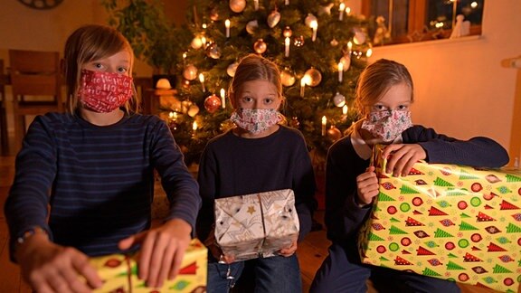 Drei Kinder mit Masken und Geschenken vor dem Weihnachtsbaum