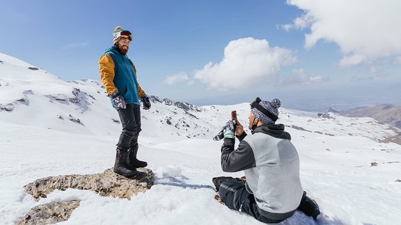 Sportler in warmer Kleidung, der im Winter ein Foto auf dem Handy auf einem Bergrücken unter bewölktem blauem Himmel macht