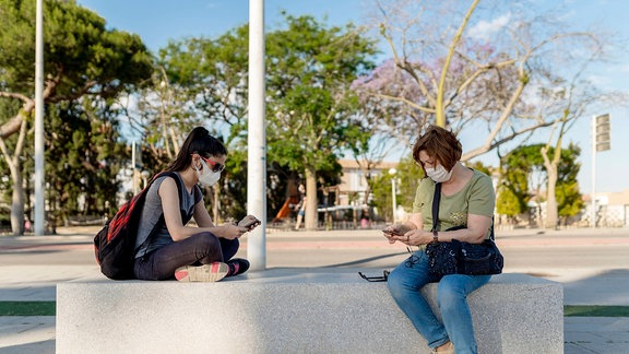 Zwei Frauen mit Mundschutz nutzen ihr Smartphone auf einer Bank sitzend (Symbolfoto)